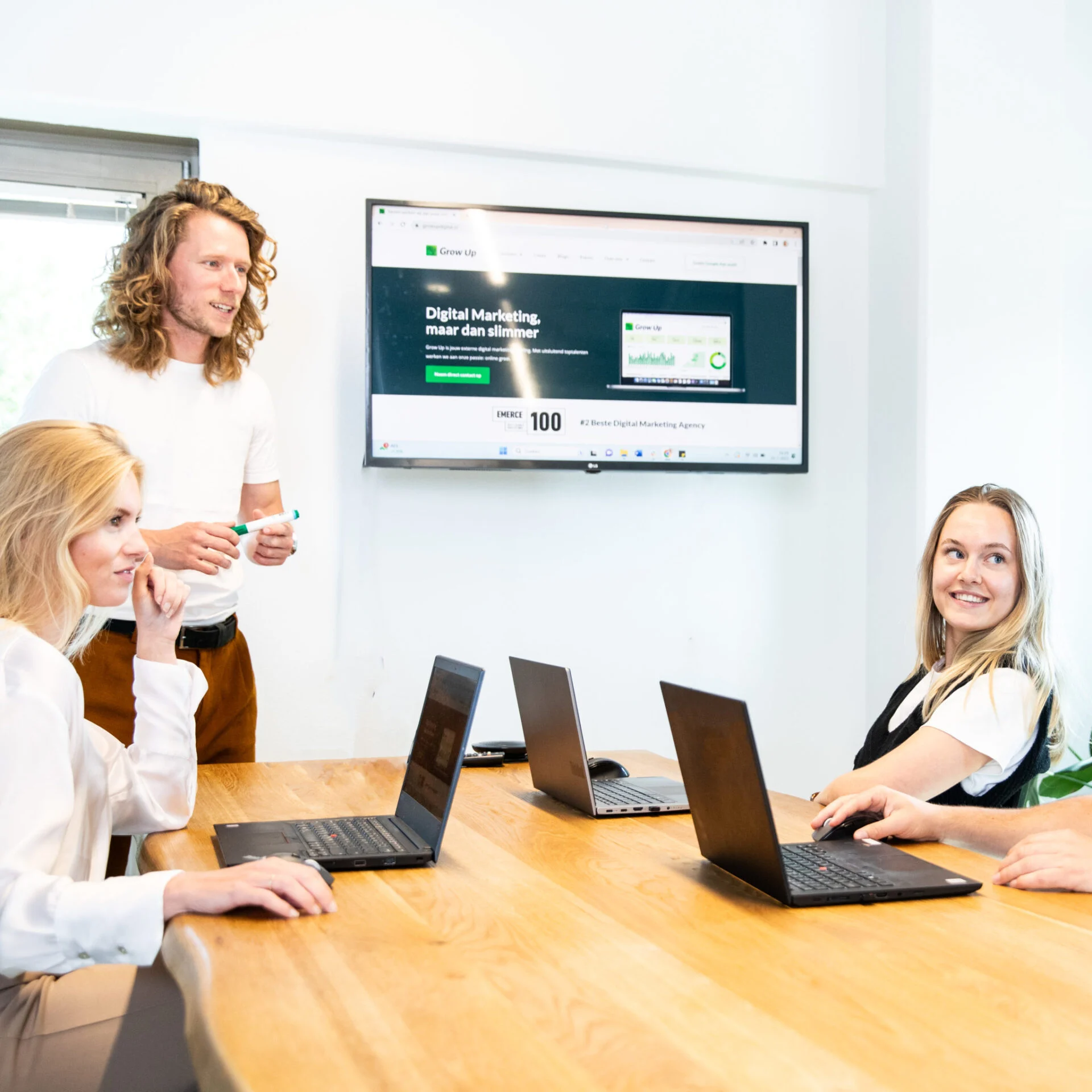 Vier mensen zitten aan een vergadertafel met open laptops, terwijl een man met lang haar voor een scherm staat en presenteert. Op het scherm staat de homepage van Grow Up Digital met de tekst “Digital Marketing, maar dan slimmer.” De sfeer is professioneel en interactief.