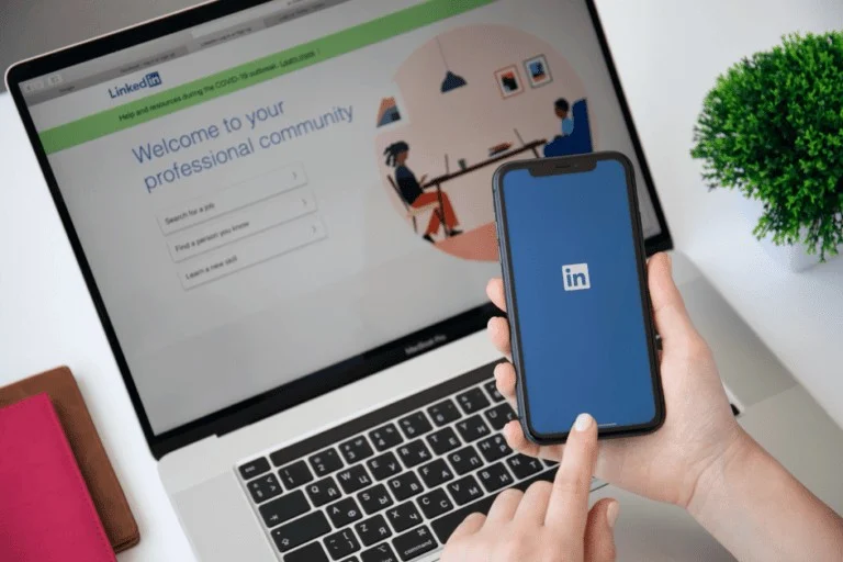 Een hand houdt een smartphone vast met het LinkedIn-opstartscherm in beeld. Op de achtergrond staat een opengeklapte laptop met een LinkedIn-loginpagina waarop staat: “Welcome to your professional community.”