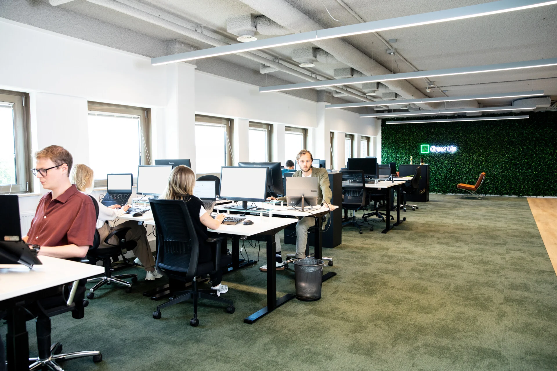 Kantoorruimte met meerdere mensen die werken aan bureaus met computerschermen. Het interieur is licht en ruimtelijk met veel ramen. Achterin is een groene muur met het verlichte logo “Grow Up Digital”.