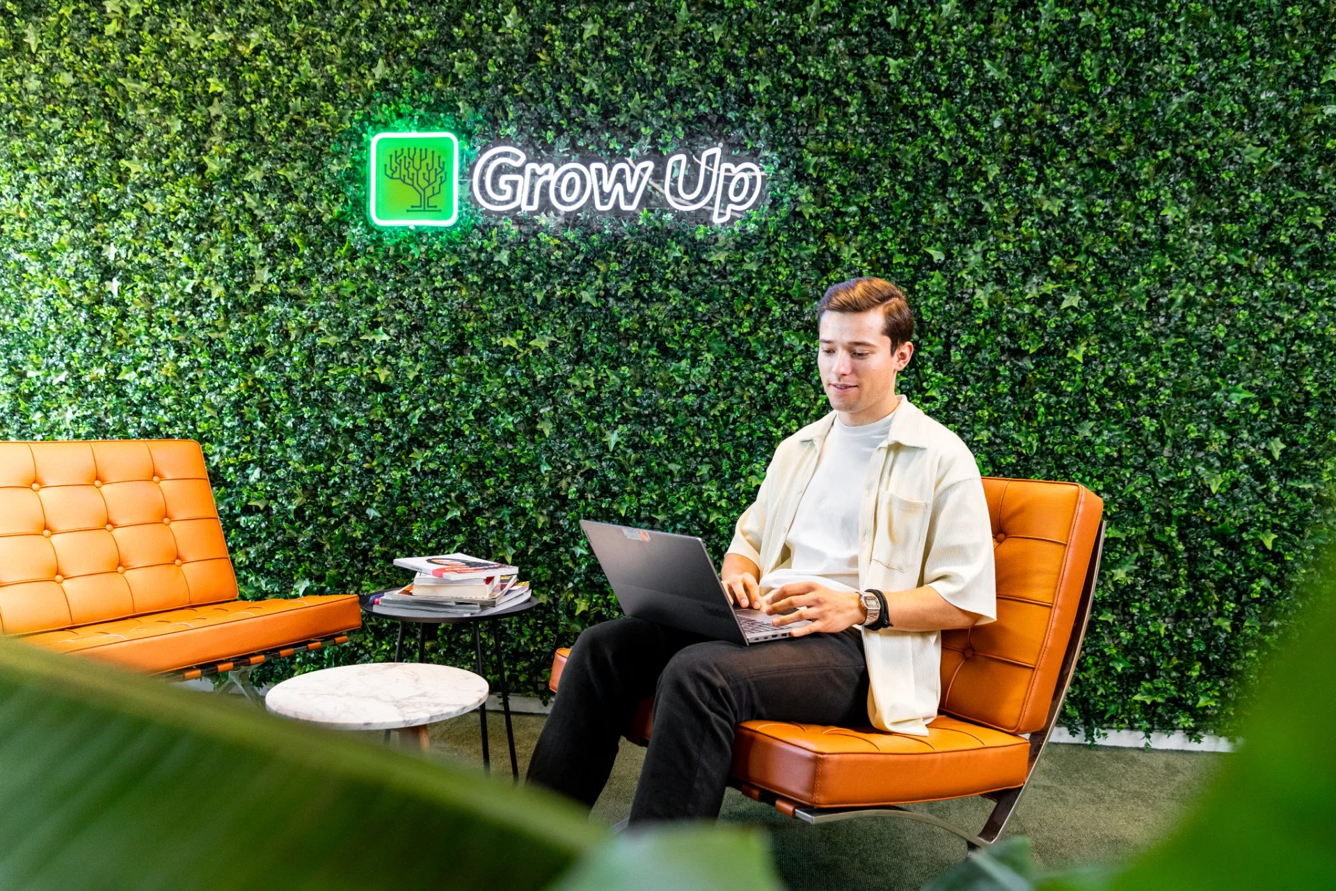 Michiel Pappenheim zit met een laptop op schoot in een oranje stoel. Hij kijkt glimlachend naar de camera. Op de achtergrond een groene plantenmuur met het verlichte logo “Grow Up Digital”.