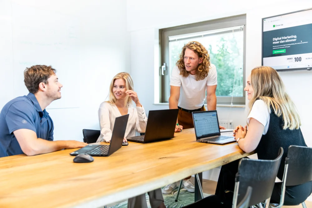 Vier collega’s overleggen aan een houten vergadertafel met open laptops. Eén man staat en spreekt, de anderen zitten en luisteren. In de achtergrond een raam met uitzicht op bomen.