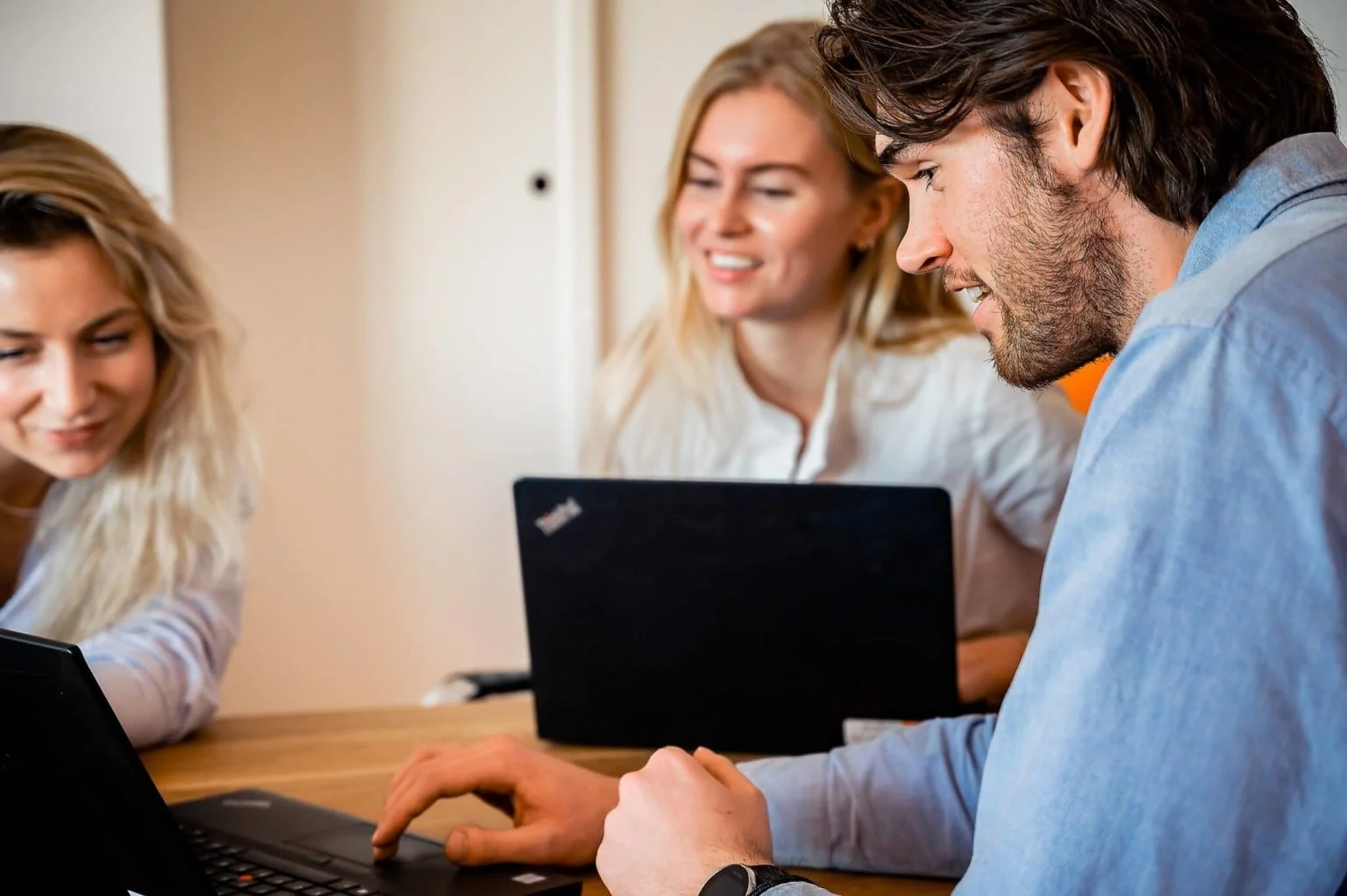 Drie collega’s van Grow Up Digital zitten dicht bij elkaar aan een tafel, lachend en geconcentreerd kijkend naar een laptopscherm. Ze dragen zakelijke kleding en werken samen in een lichte kantoorruimte.